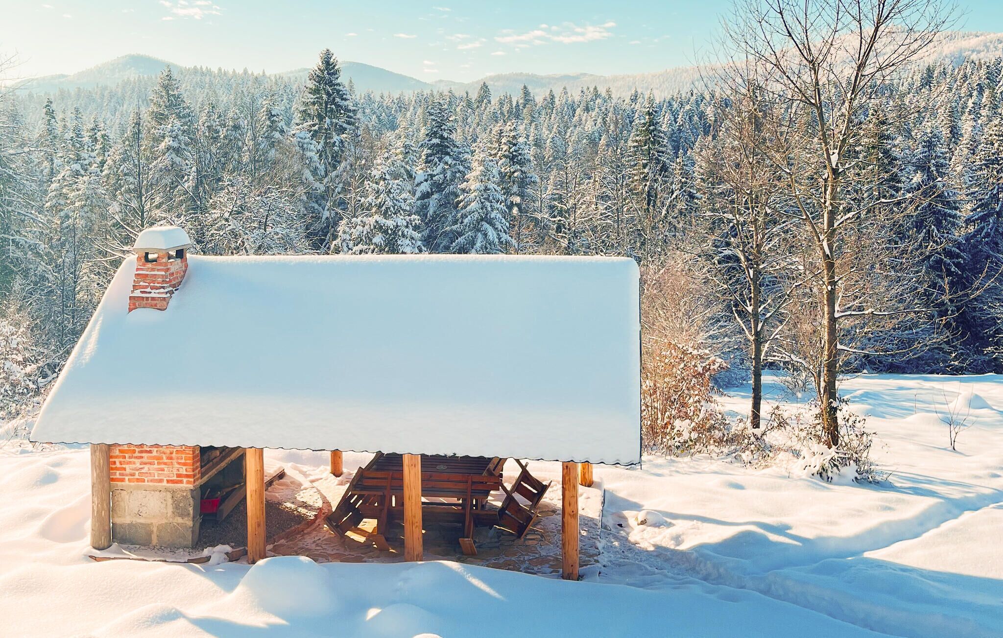 Une cabane en bois recouverte de neige, avec une cheminée en briques, entourée d’un paysage enneigé et d’une forêt dense.