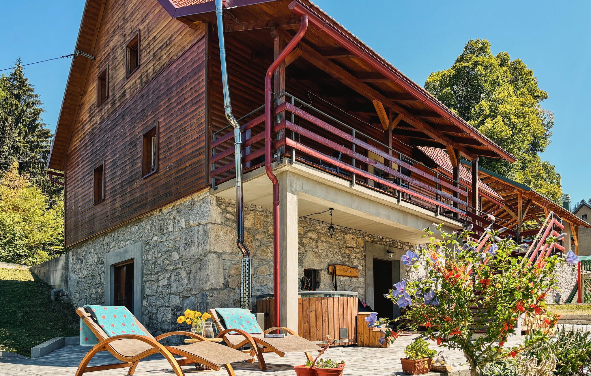 Edificio de madera con balcón, base de piedra y zona de estar al aire libre con sillas y plantas en macetas.