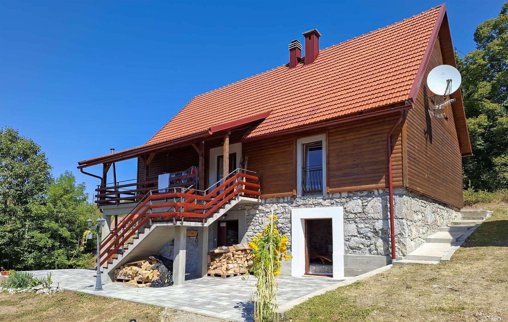 A two-story wooden house with a red-tiled roof, a satellite dish, and a stone foundation.