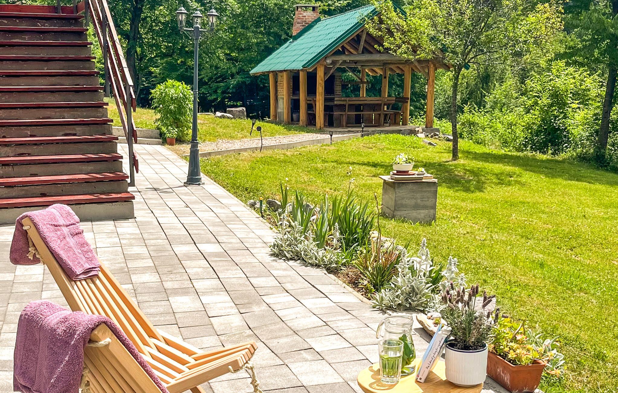 A wooden bench with a towel, a stone pathway, a gazebo, and a grassy area with potted plants.