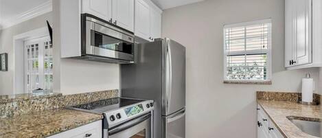 Una cocina con armarios blancos, electrodomésticos de acero inoxidable, encimeras de granito y una ventana con persianas.