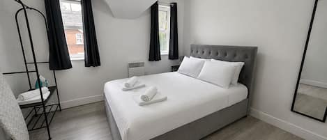 Ein modernes Schlafzimmer mit einem großen Bett, einem Standheizkörper, einem Spiegel und einem Fenster mit Vorhängen.