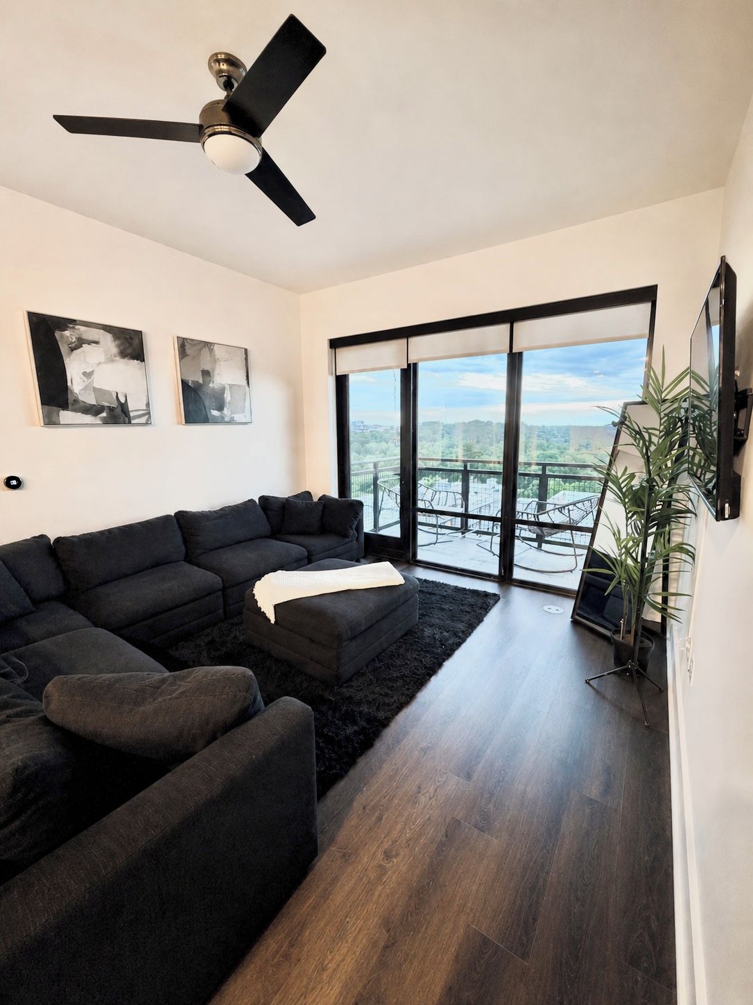 A modern living room with a sectional sofa, a ceiling fan, and a large window offering a view of a balcony.