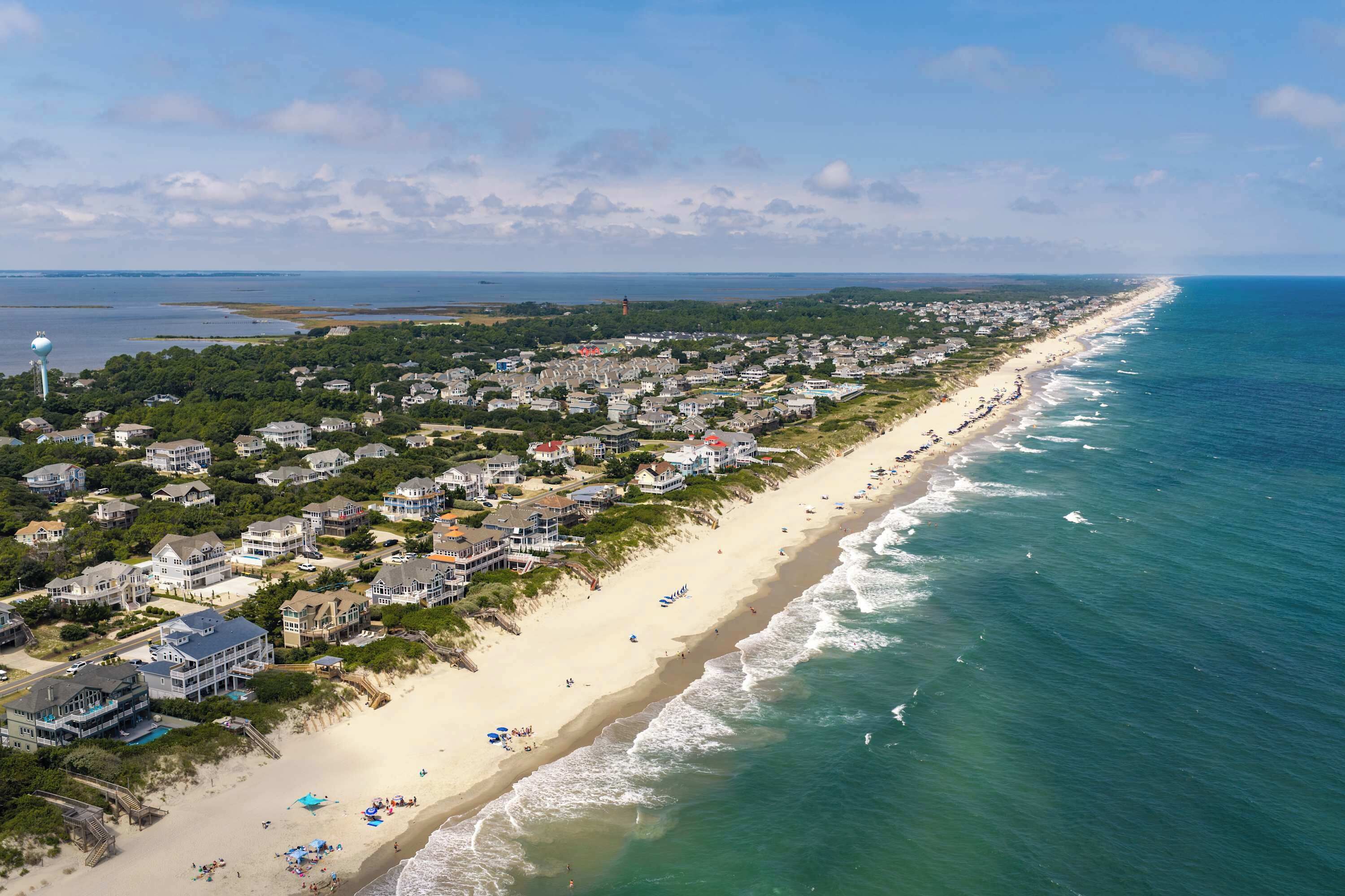 An aerial view of a coastal residential area with houses, a beach, and a lighthouse.
