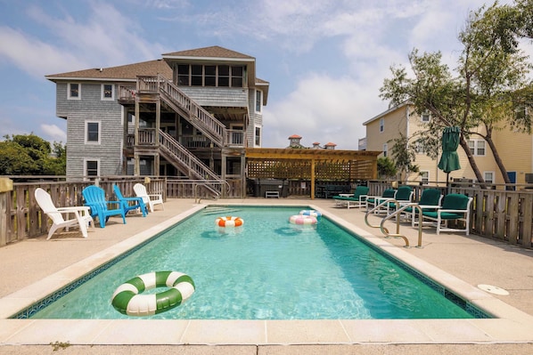 A swimming pool with lounge chairs and inflatable rings, surrounded by a wooden deck and a multi-story building.