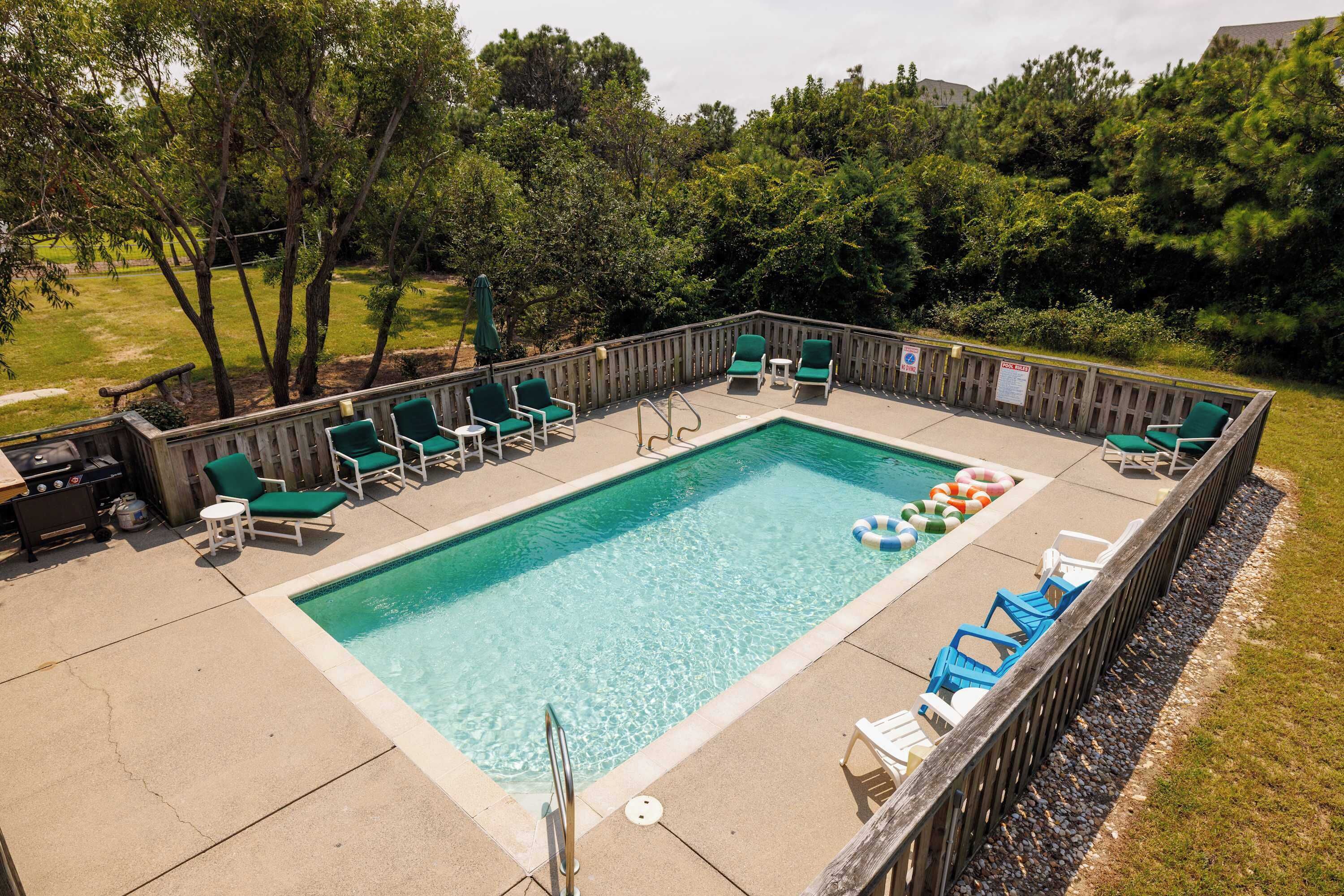 A rectangular outdoor swimming pool with a wooden deck, surrounded by green chairs and a wooden fence.