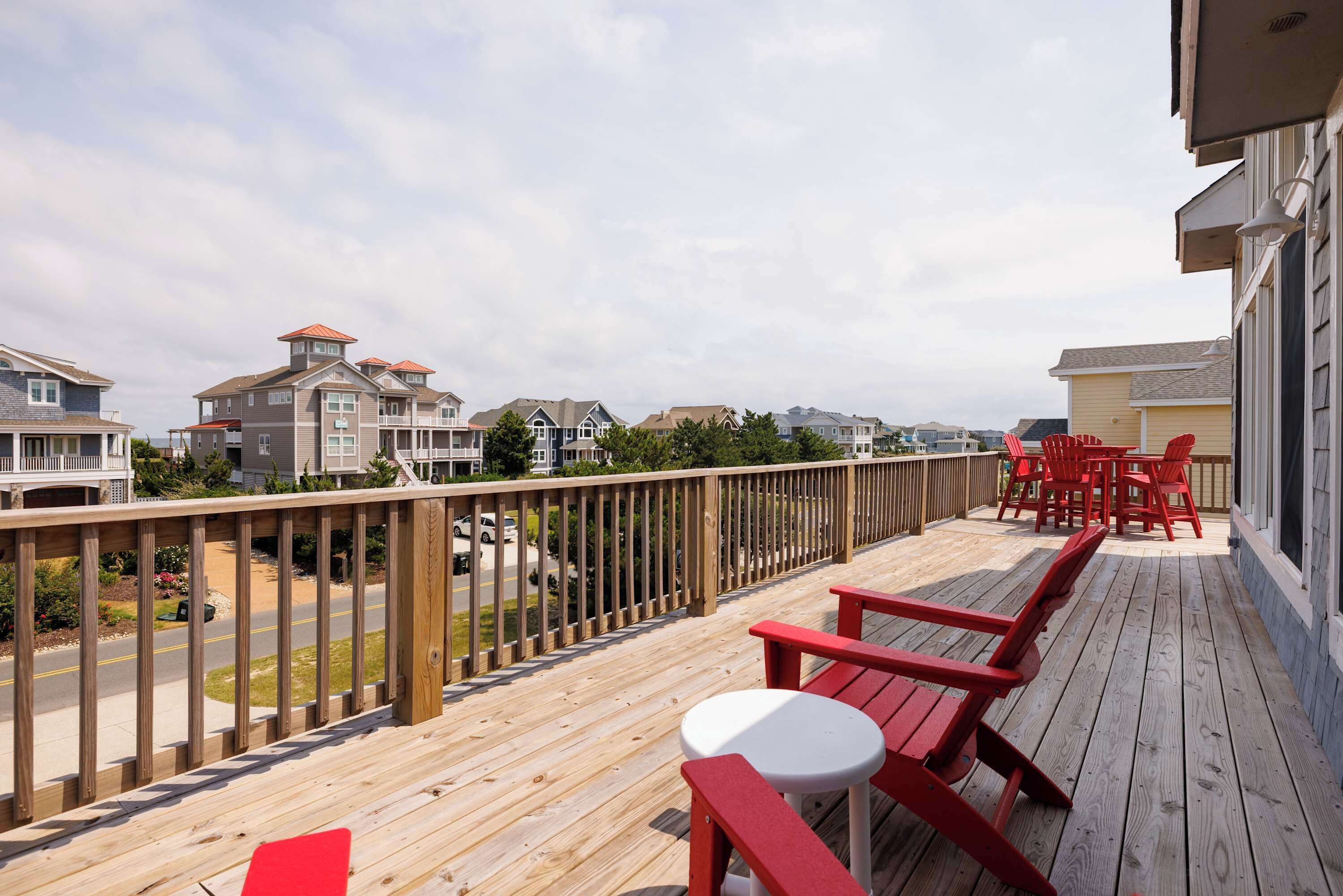 A wooden deck with red chairs and a white table, overlooking a residential area with houses and a road.