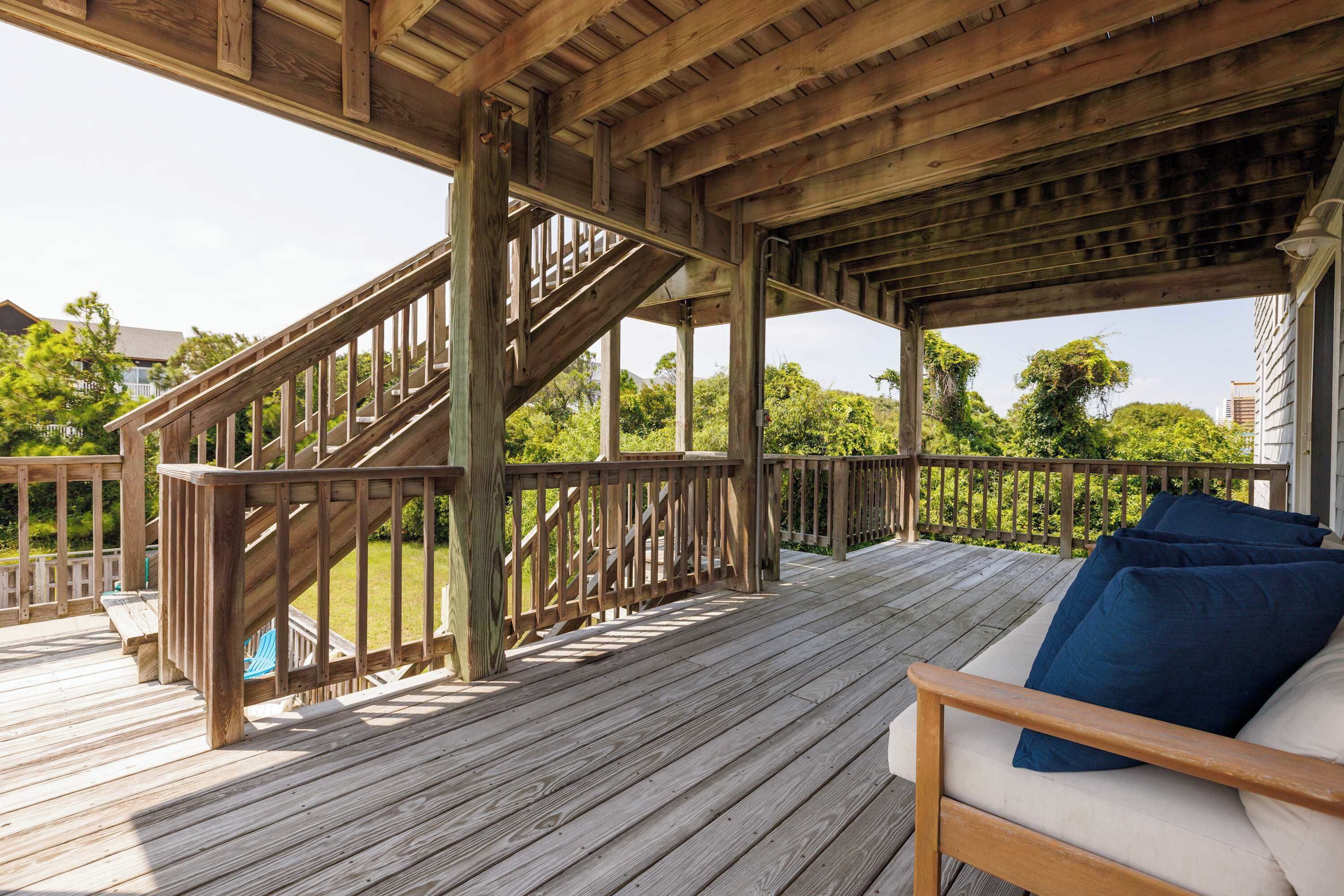 Une terrasse en bois avec un escalier menant à un niveau supérieur, un coin salon avec des coussins bleus, et une vue sur la verdure et les bâtiments en arrière-plan.