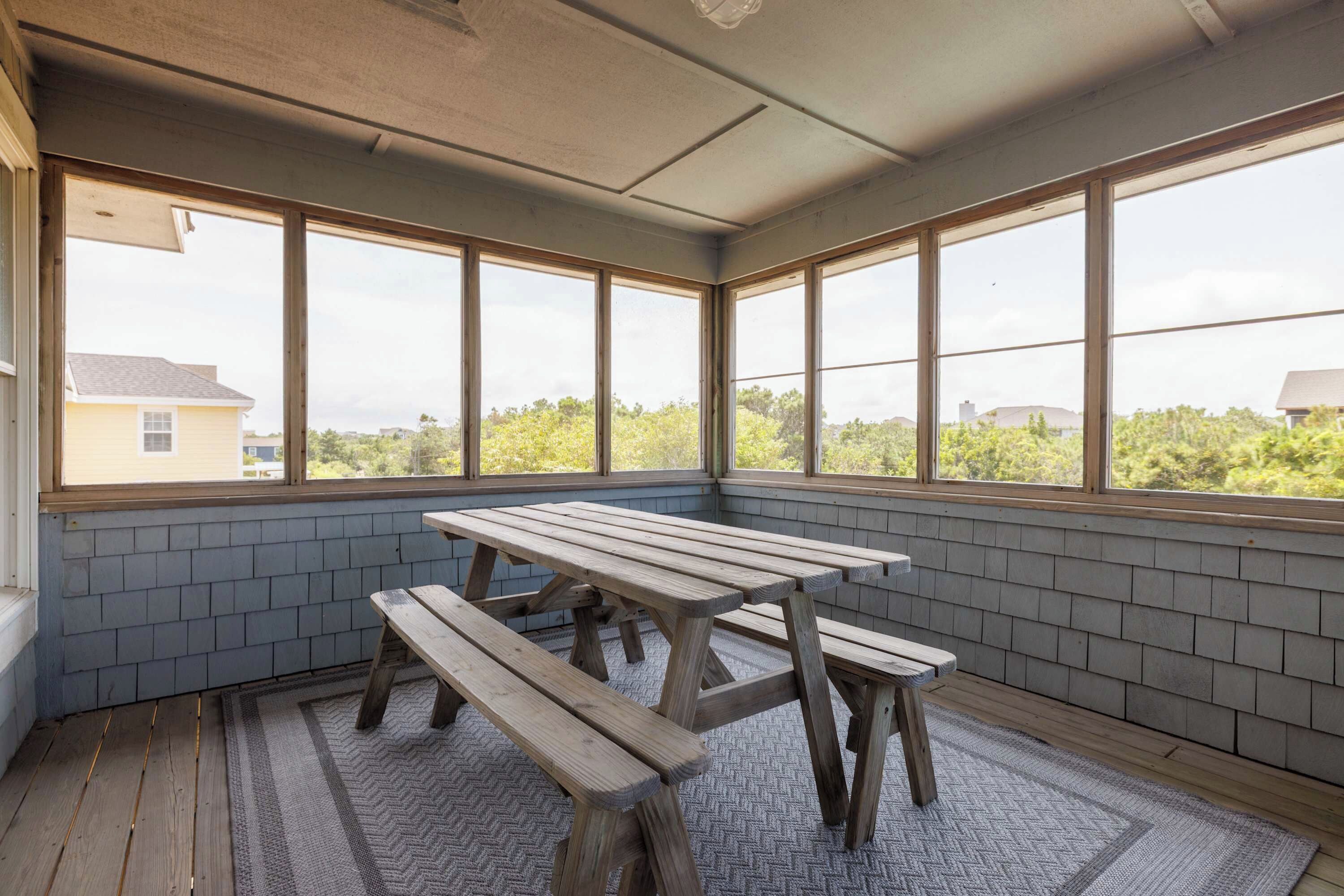 A sunroom with a wooden table and benches, large windows, and a view of greenery and buildings.