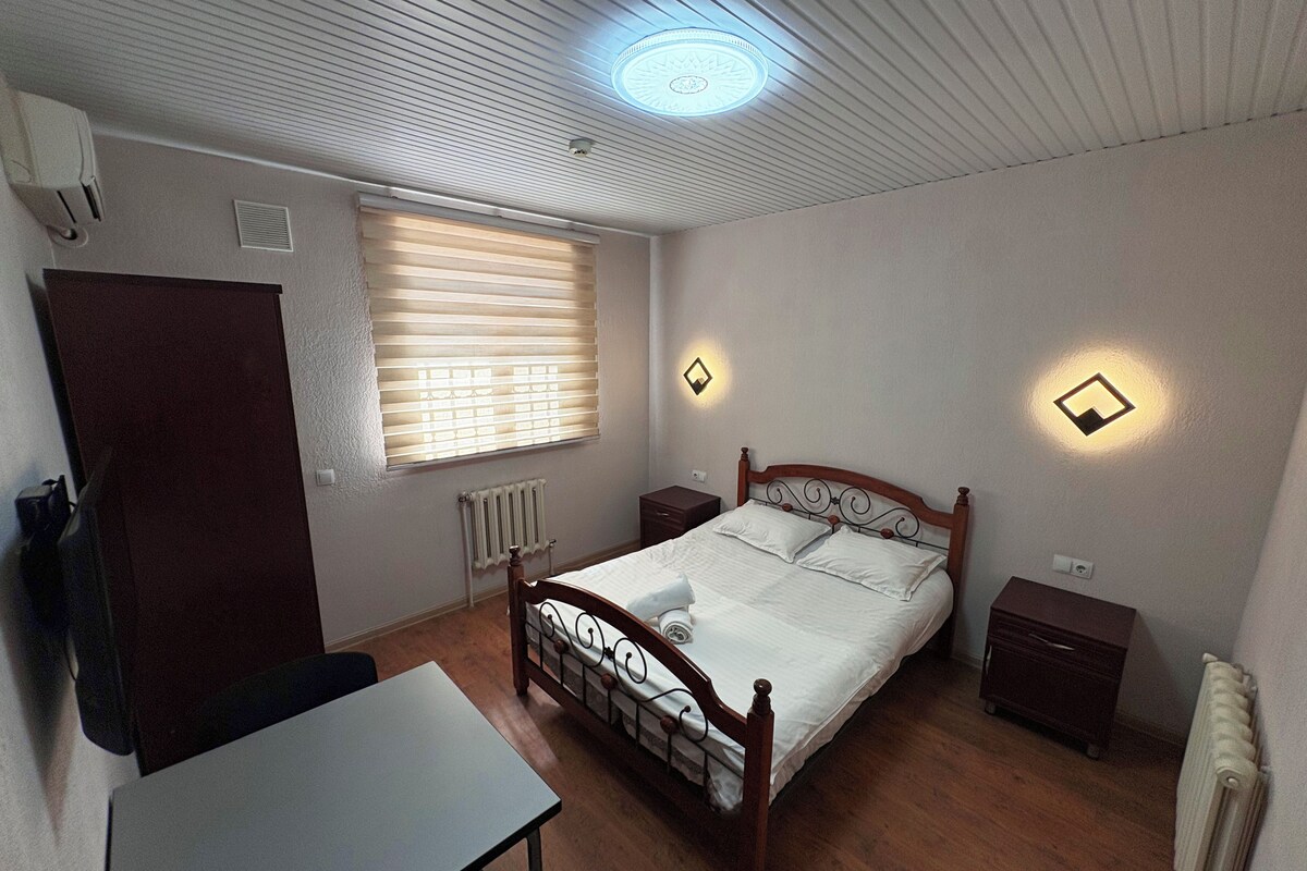Una camera da letto con un letto di legno, comodini, un termosifone, una finestra con persiane e un lampadario al soffitto.