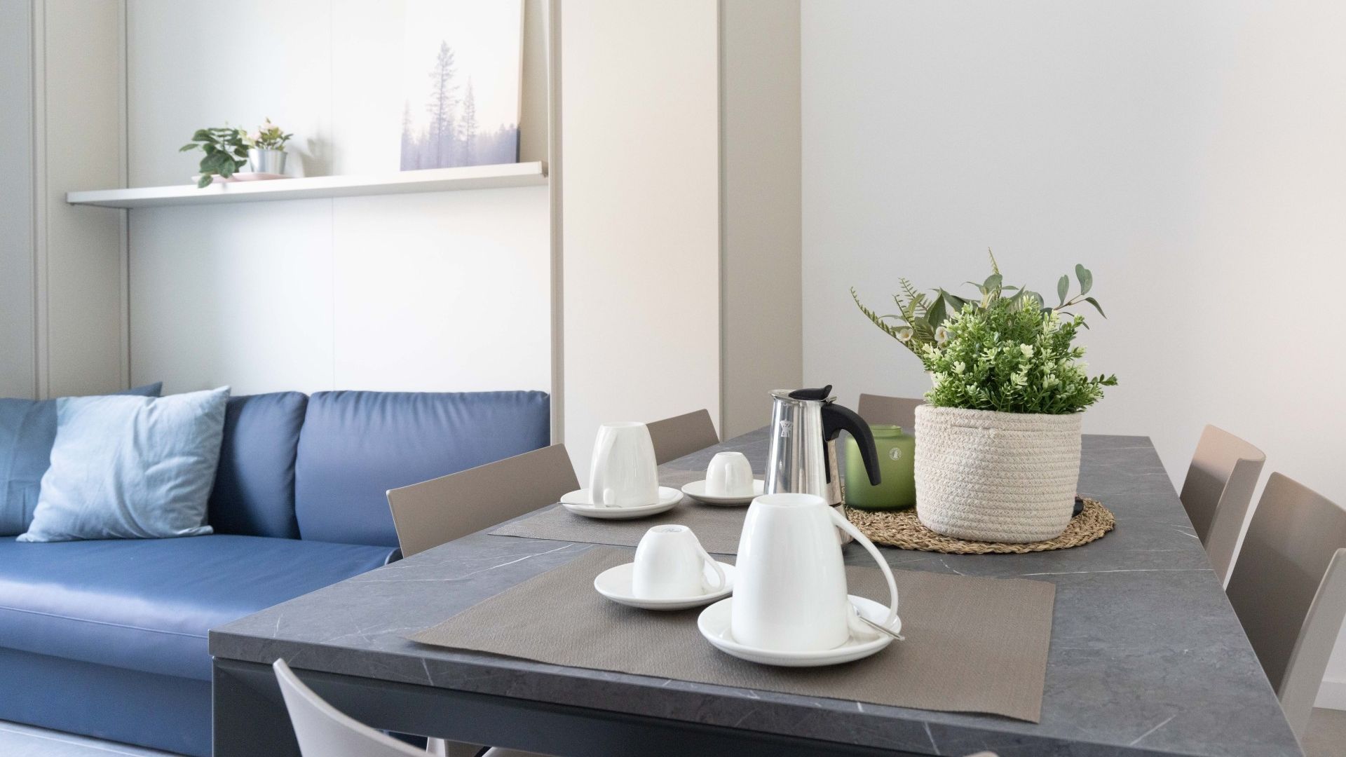 Un comedor moderno con una mesa lista para cuatro personas, un sofá azul y una planta en maceta.
