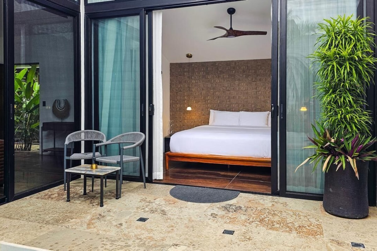 Et moderne hotelværelse med en seng, et siddeområde med stole, en potteplante og en glasdør, der fører til et udendørs område.