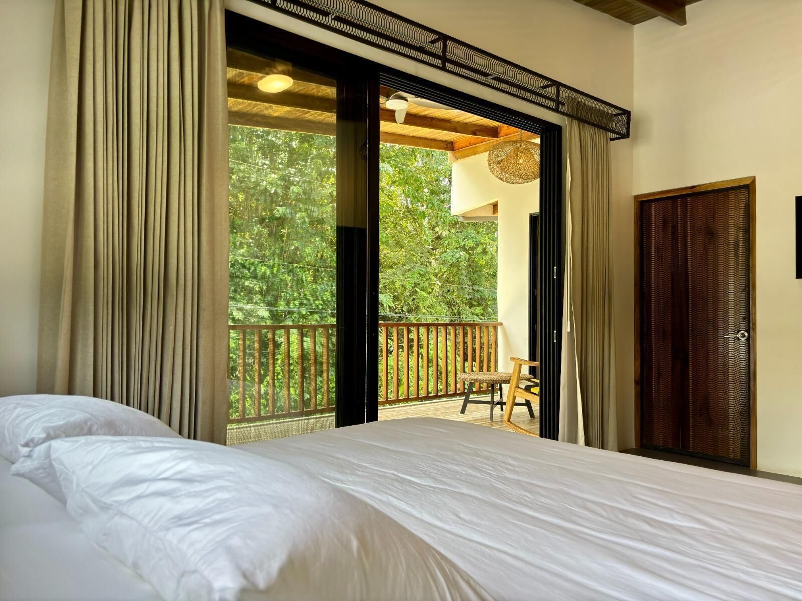 Un dormitorio con cama, un balcón con una silla y vistas al verdor exterior.