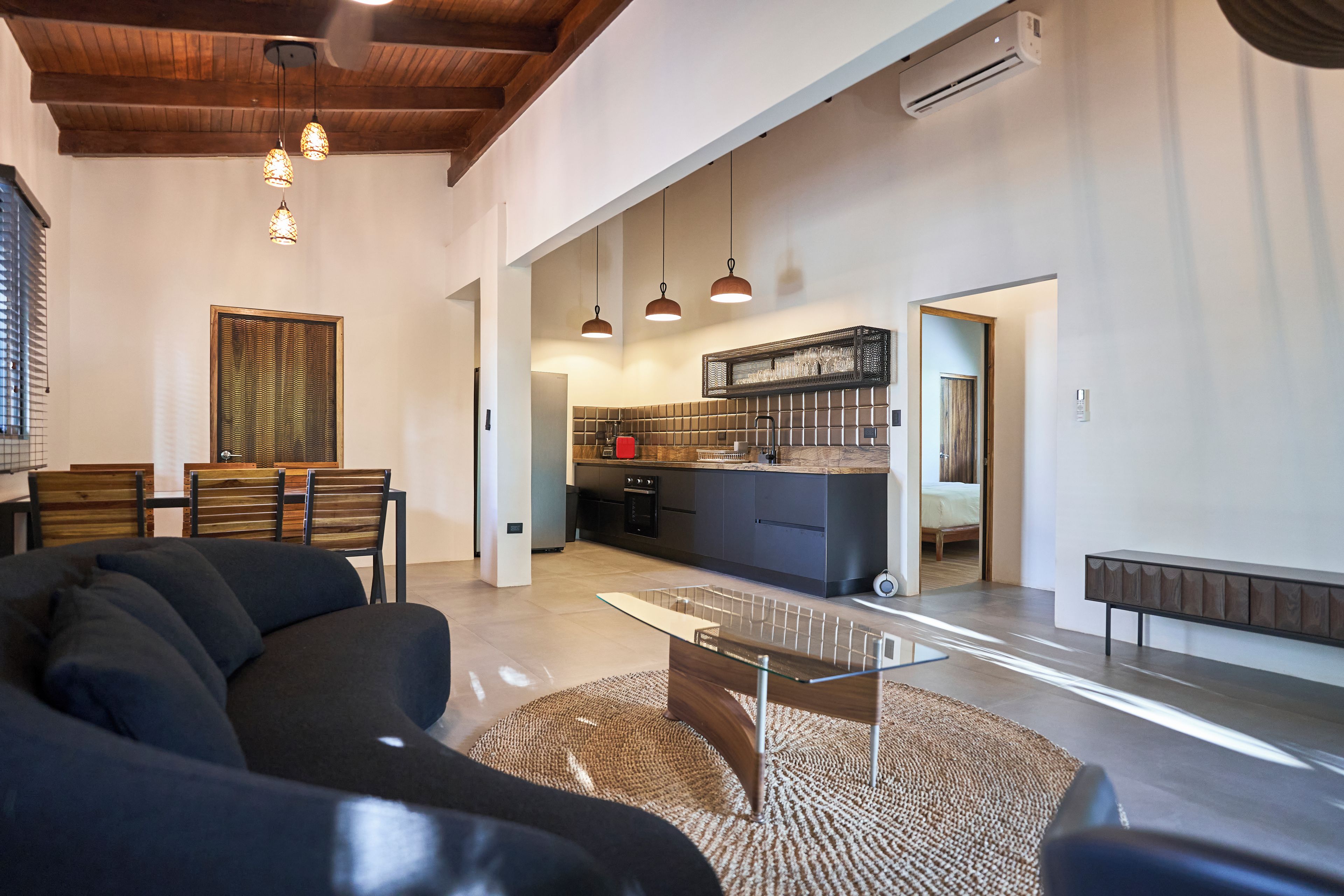 Amplia sala de estar con un sofá moderno, una mesa de centro de vidrio y un comedor. Se ve una cocina al fondo con horno empotrado y un botellero.
