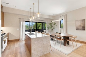 Une cuisine moderne dotée d’un îlot central, d’un coin repas avec une table et des chaises, et de grandes fenêtres offrant une vue sur la verdure.