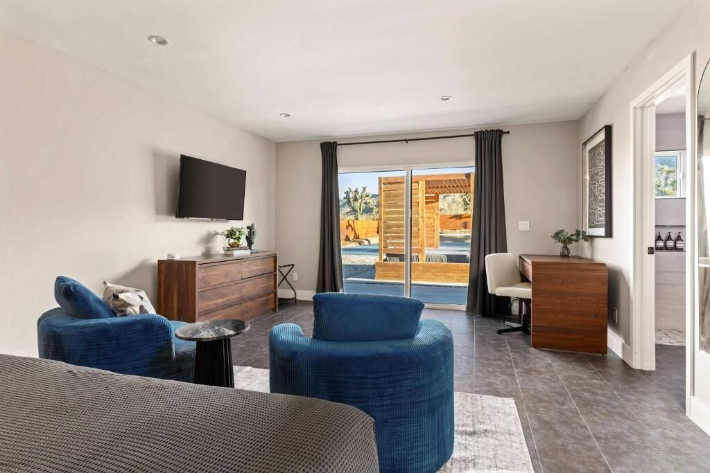 Een moderne hotelkamer met een flatscreen-tv, een houten bureau, twee blauwe fauteuils, een bank en uitzicht op het buitenzwembad.