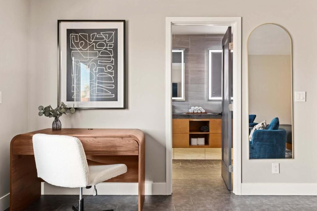 A modern room with a wooden desk, a white chair, a framed abstract artwork, a bathroom with a mirror, and a blue chair.