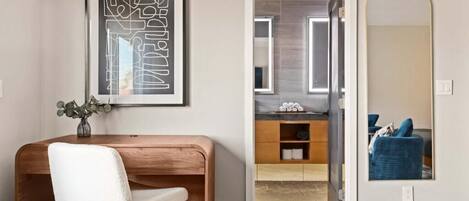 A modern room with a wooden desk, a white chair, a framed abstract artwork, a bathroom with a mirror, and a blue chair.