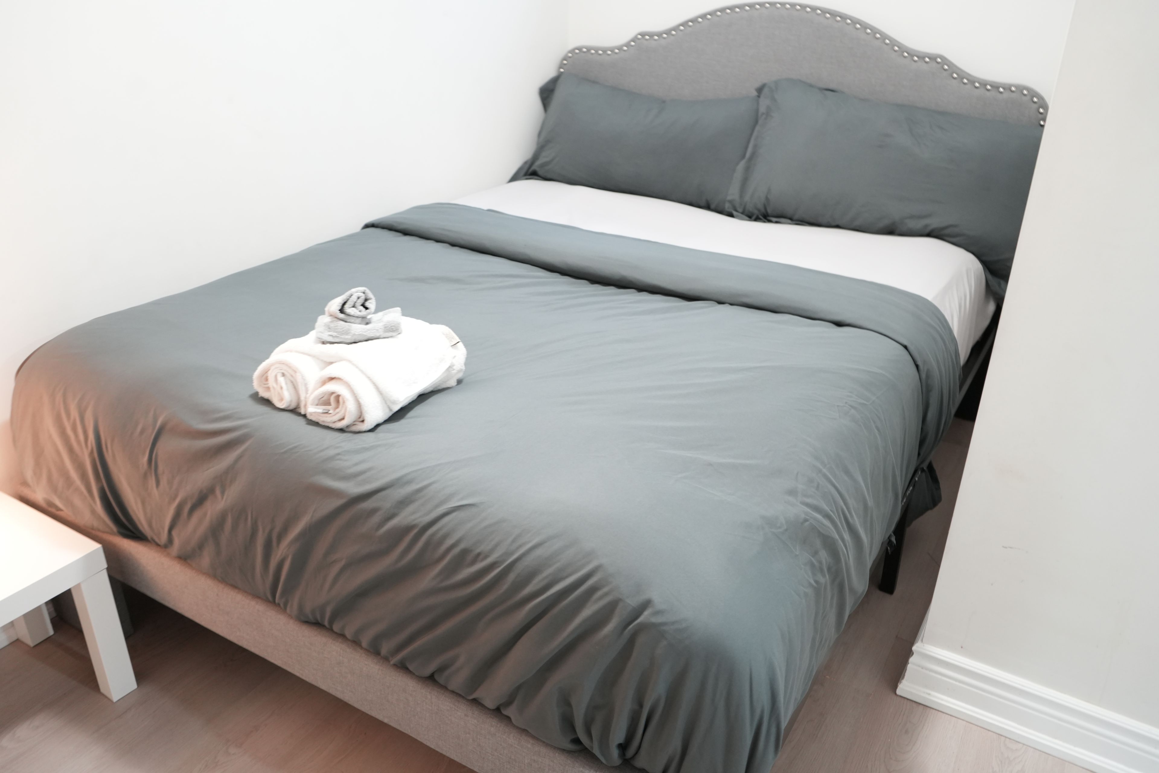 Una cama bien hecha con ropa de cama gris y una toalla blanca enrollada a la izquierda.