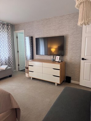 Een kamer met een wandgemonteerde flatscreen televisie, een houten dressoir met laden, een grijze bank en een kamerbreed gordijn.
