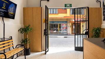 A hotel reception area with a wooden bench, potted plants, and a view of the street outside.
