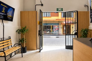 Zona de recepción de hotel con un banco de madera, plantas en macetas y vistas a la calle.