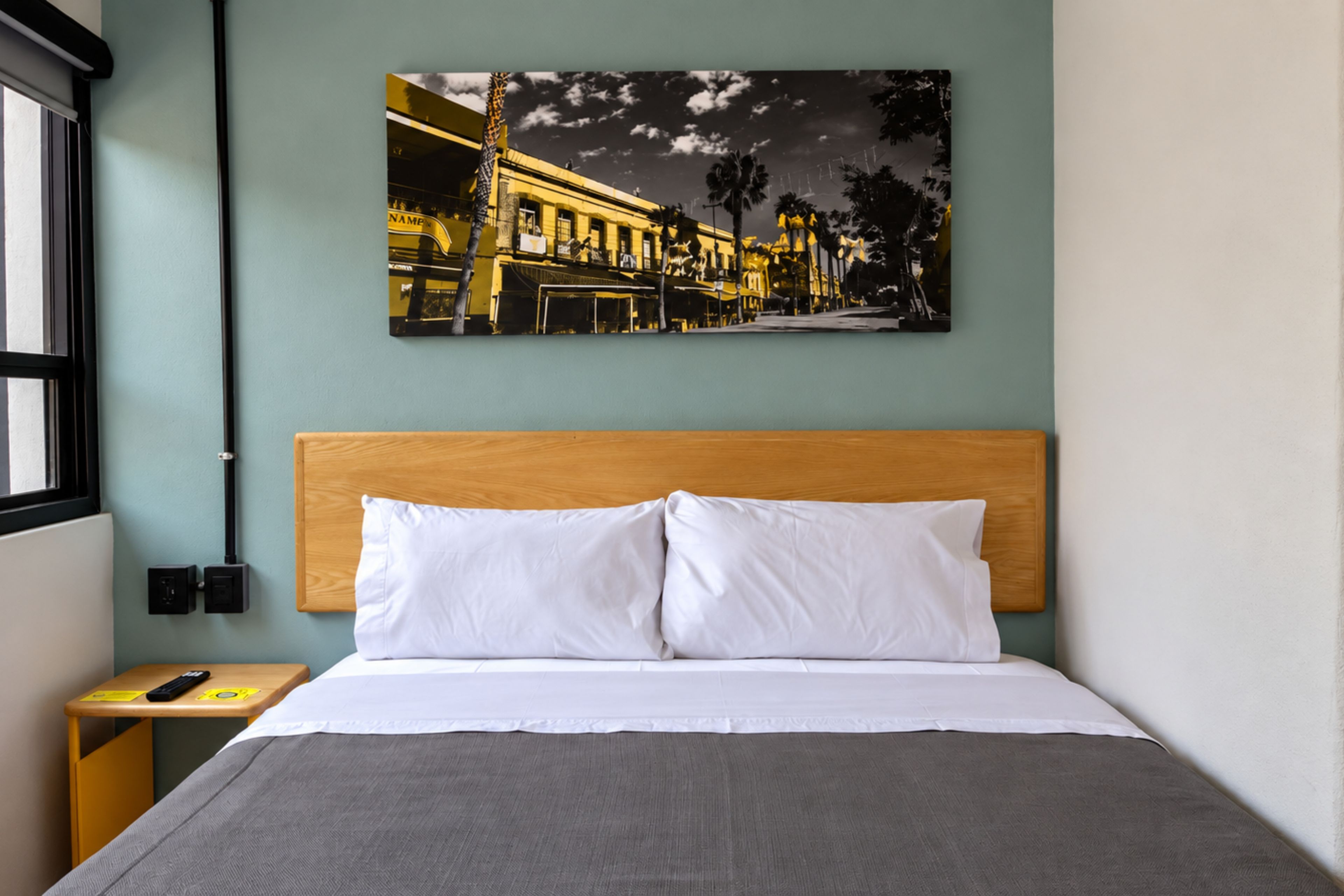 A hotel room with a bed, a nightstand, a wall-mounted picture, and a window.