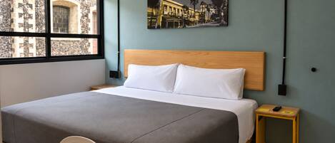 A modern hotel room with a large bed, a small bedside table, a chair, a window with a view of a stone building, and a wall-mounted artwork.