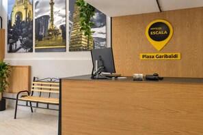 Un comptoir d’accueil d’hôtel avec un ordinateur, une enseigne indiquant « HOTELES ESCALA Plaza Garibaldi », et une grande fenêtre donnant sur le paysage urbain.