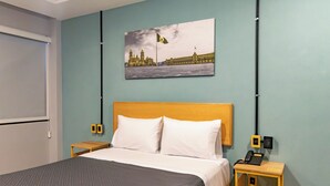 Une chambre d’hôtel avec un lit, une table de chevet avec un téléphone, un interrupteur fixé au mur et un tableau représentant un bâtiment.