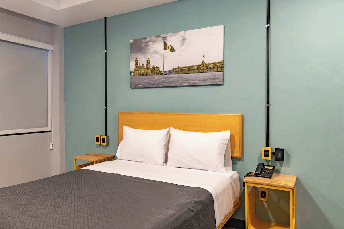 Una camera d'albergo con un letto, un comodino con un telefono, un interruttore della luce montato a parete e un quadro raffigurante un edificio.