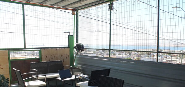A room with a large window overlooking a cityscape, a table with chairs, and a hanging plant. - Disegni 22 · Caldera Family Department (Caldera)