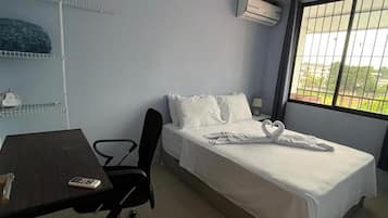 Una cama individual con ropa de cama blanca, un escritorio con silla y una ventana con vista a follaje.