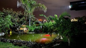 Un giardino notturno con uno stagno, palme e un edificio sullo sfondo.
