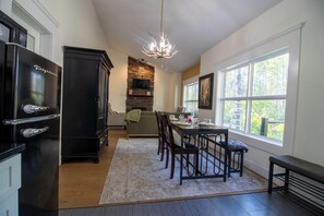 A dining area with a table set for four, a black refrigerator, a stone fireplace, and a large window with a view of trees.