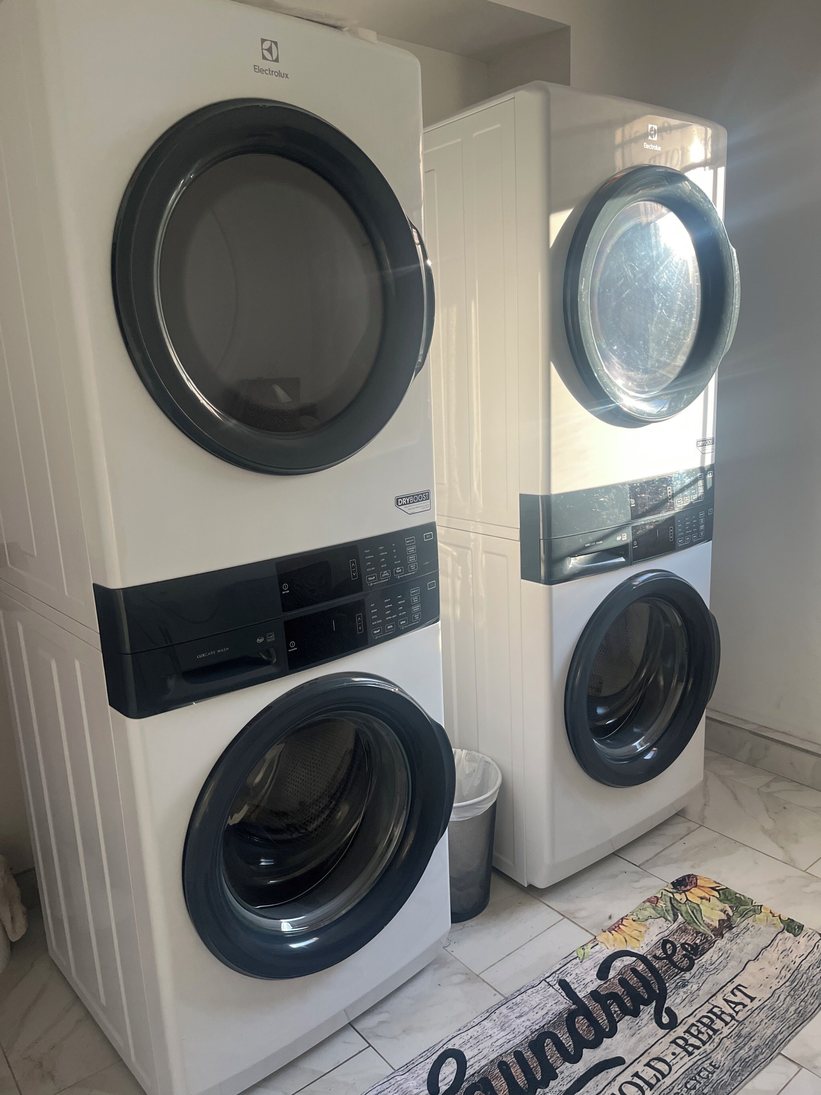 Deux machines à laver à chargement frontal se trouvent dans une buanderie dont le sol est carrelé et où est posé un tapis orné de motifs textuels.
