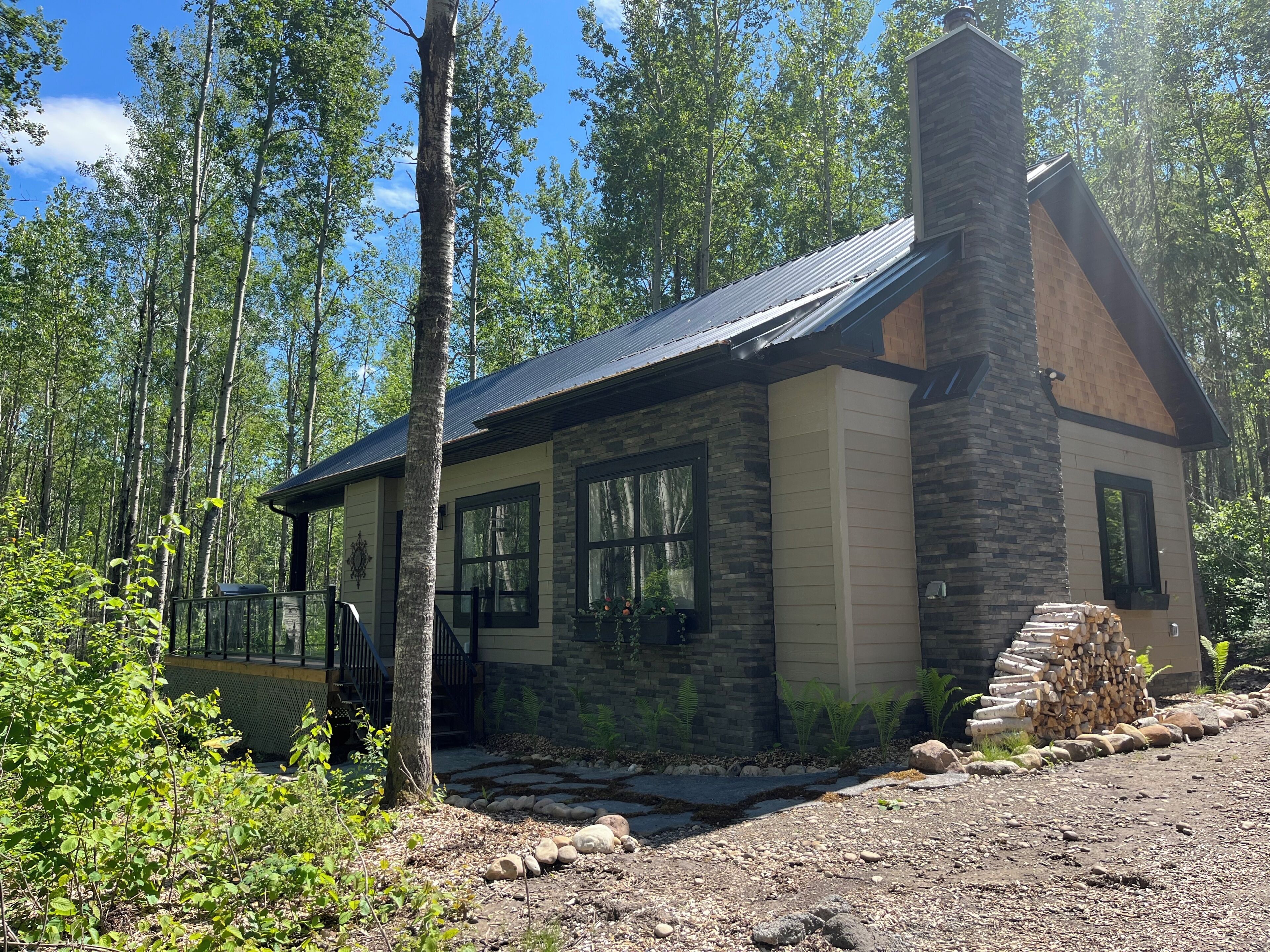Une cabane dotée d’une cheminée en pierre, de grandes fenêtres et d’une terrasse en bois.