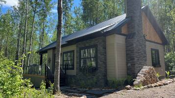 Une cabane dotée d’une cheminée en pierre, de grandes fenêtres et d’une terrasse en bois.