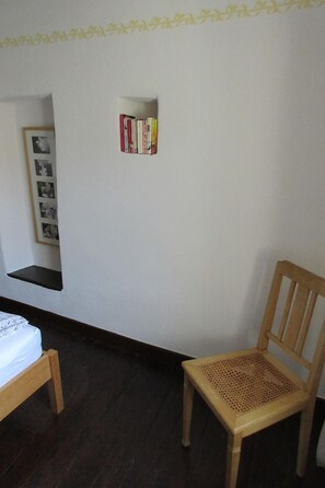 Ein Zimmer mit einem Bett, einem Holzstuhl, einem Regal mit Büchern und einem gerahmten Foto.