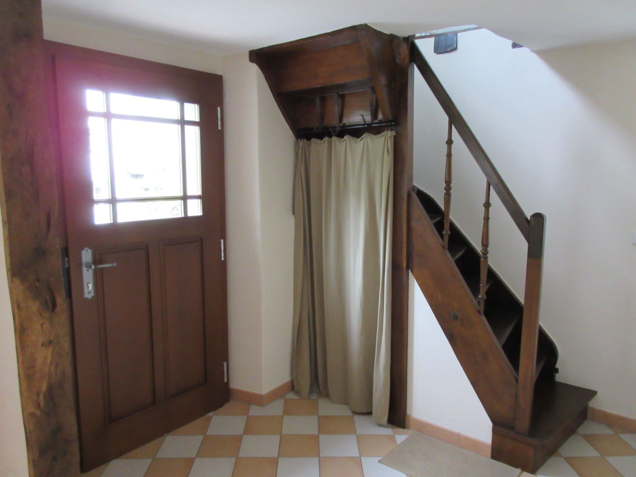 Una habitación con puerta de madera, ventana con vistas, escalera con barandilla de madera y suelo de baldosas.