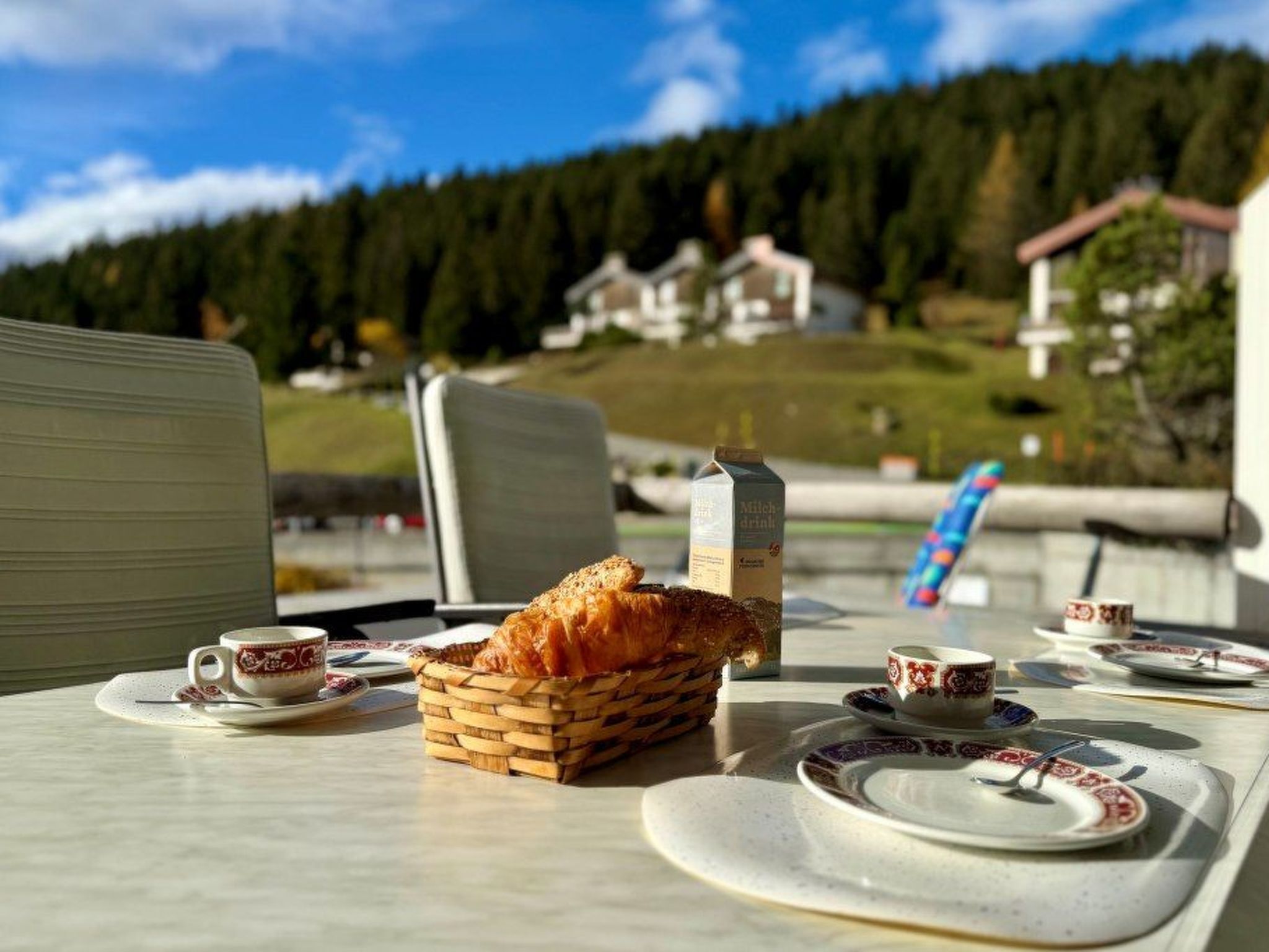 Una mesa servida con una cesta de pan, una botella y platos con tazas, con vistas a un paisaje montañoso.