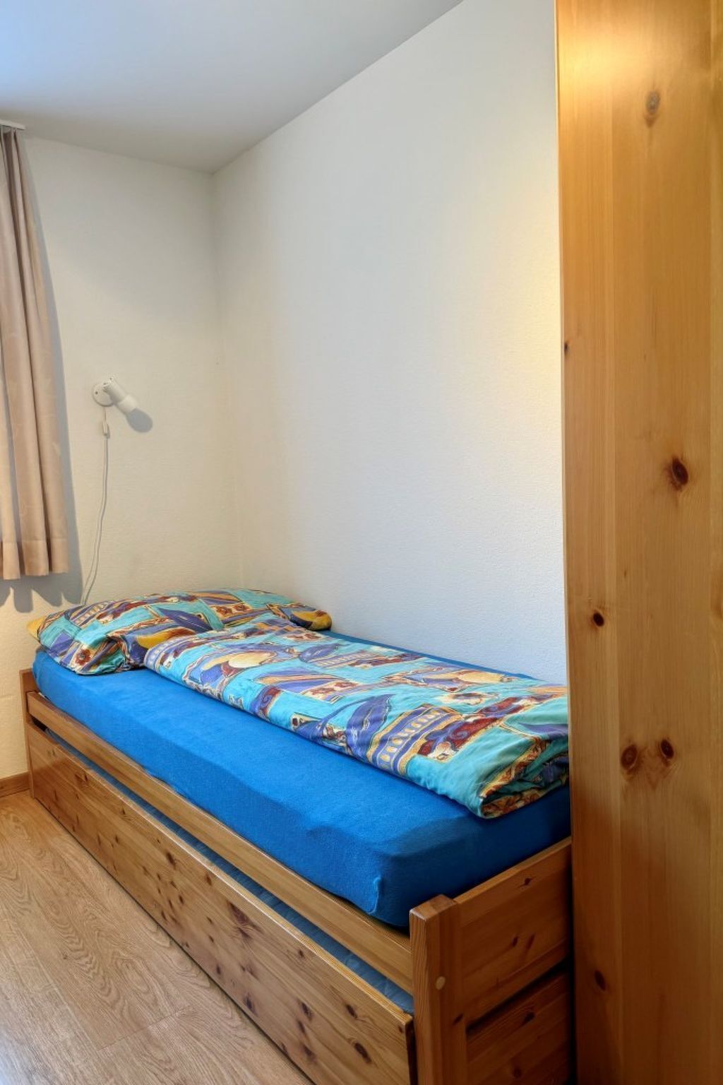 A small, single-bed room with a wooden frame, a blue mattress, and a colorful blanket.