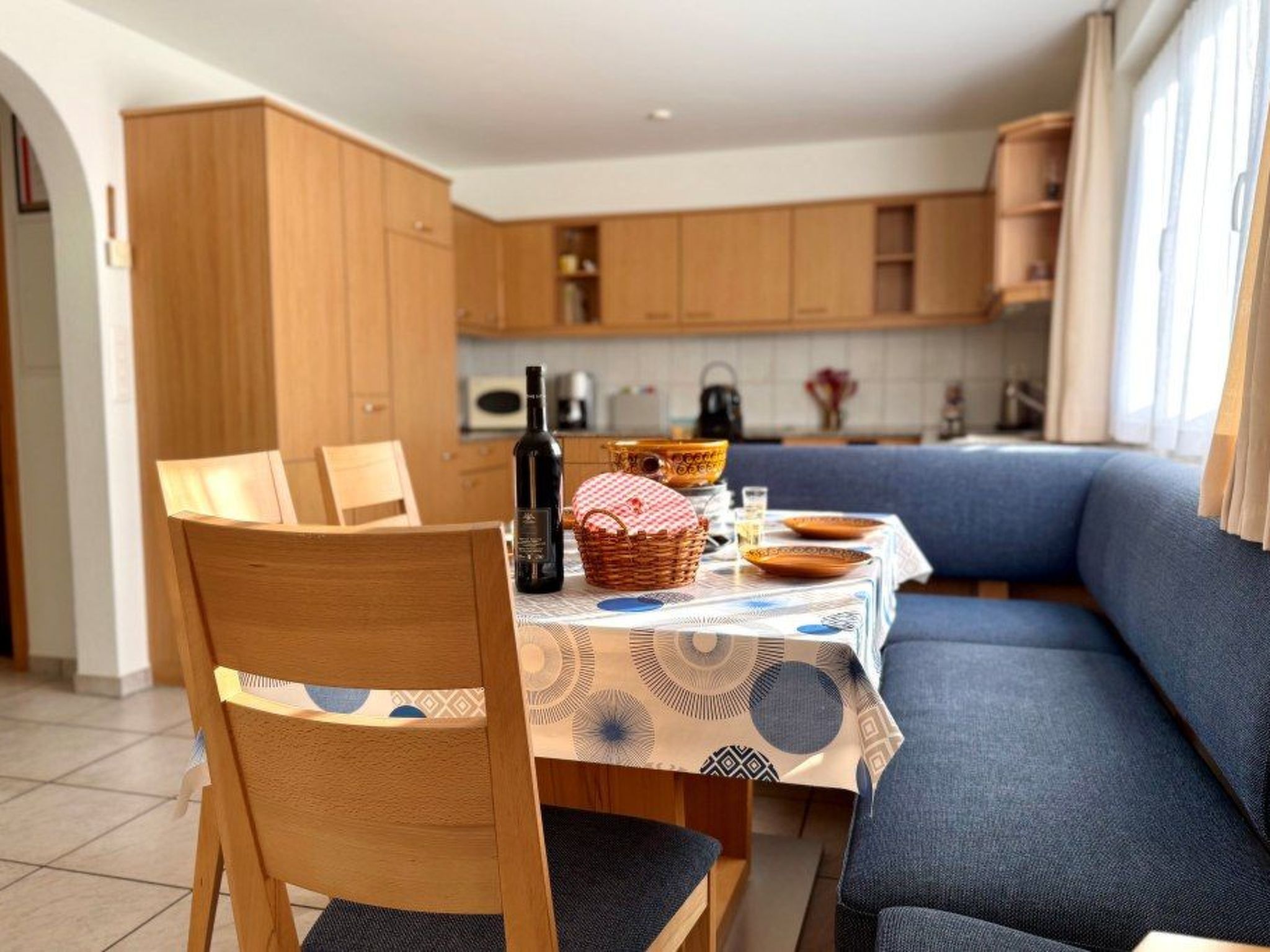 Un coin repas avec des chaises en bois et une table dressée pour deux personnes, un canapé bleu et, au fond, une cuisine.