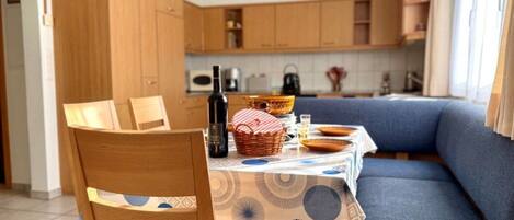 Un coin repas avec des chaises en bois et une table dressée pour deux personnes, un canapé bleu et, au fond, une cuisine.