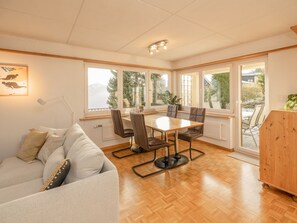 A room with a wooden floor, a dining table with chairs, a sofa, and large windows with a view of greenery and mountains.