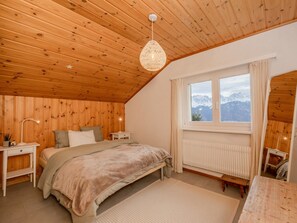A cozy bedroom with a wooden ceiling, a large bed, a bedside table, a nightstand, a hanging light fixture, a window with a view of mountains, and a radiator.