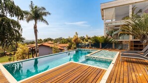 A swimming pool with wooden deck, surrounded by palm trees and houses, under a clear sky. - Casa em Condom nio com Piscina e Sinuca em Itupeva (Itupeva)
