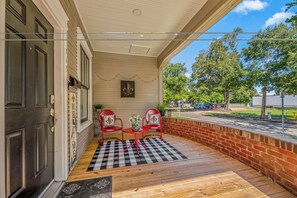 A covered porch with two red chairs and a table, a black and white checkered mat, and a brick wall.