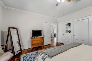A bedroom with a large mirror, a flat-screen TV, a wooden dresser, a bed with a grey blanket, and a hallway leading to another room.