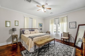 A bedroom with a bed, bedside tables, a bench, a chair, a ceiling fan, and a large window with curtains.